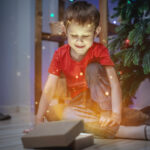 Happy little boy is sitting next to a Christmas tree and opening a gift box from which a magical light and shine. Concept of New Year mood and festive atmosphere